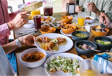 Grupo de amigos comiendo tacos mexicanos y comida tradicional, bocadillos y con las manos sobre la mesa, vista aérea. Cocina mexicana. 