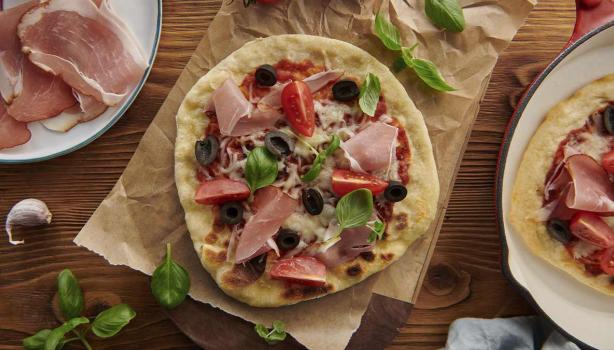 Mesa con pizza de prosciutto con aceitunas, tomates y arúgula