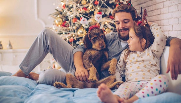 Padre y su adorable hija recostados en la cama con su perro. Al fondo, árbol de Navidad. La niña lleva una diadema con cuerno