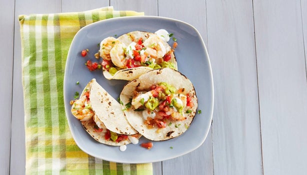 Tacos con cebolla, cilantro, guacamole y camarones en plato azul sobre servilleta verde sobre mesa de madera.