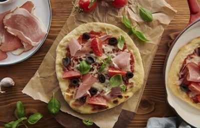 Mesa con pizza de prosciutto con aceitunas, tomates y arúgula