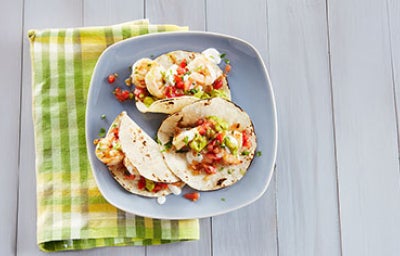 Tacos con cebolla, cilantro, guacamole y camarones en plato azul sobre servilleta verde sobre mesa de madera.
