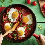 Huevos shakshuka con hierbas, salsa de tomate, pan y pimentón picado en toma cenital.