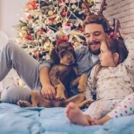Padre y su adorable hija recostados en la cama con su perro. Al fondo, árbol de Navidad.