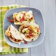Tacos con cebolla, cilantro, guacamole y camarones en plato azul sobre servilleta verde sobre mesa de madera.