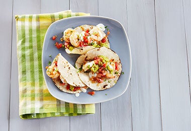 Tacos con cebolla, cilantro, guacamole y camarones. Tacos con cebolla, cilantro, guacamole y camarones en plato azul sobre servilleta verde en mesa de madera.